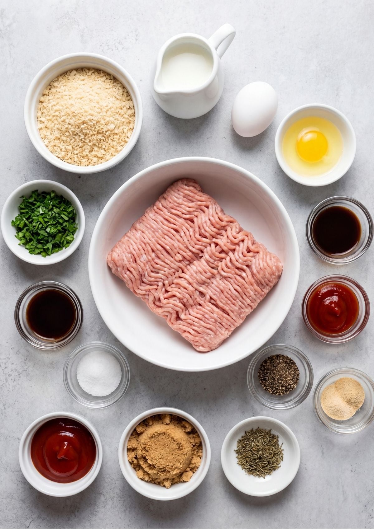 Overhead flat lay of recipe ingredients including ground turkey, panko breadcrumbs, milk, eggs, chopped parsley, brown sugar, ketchup, and spices in small white bowls on a gray counter