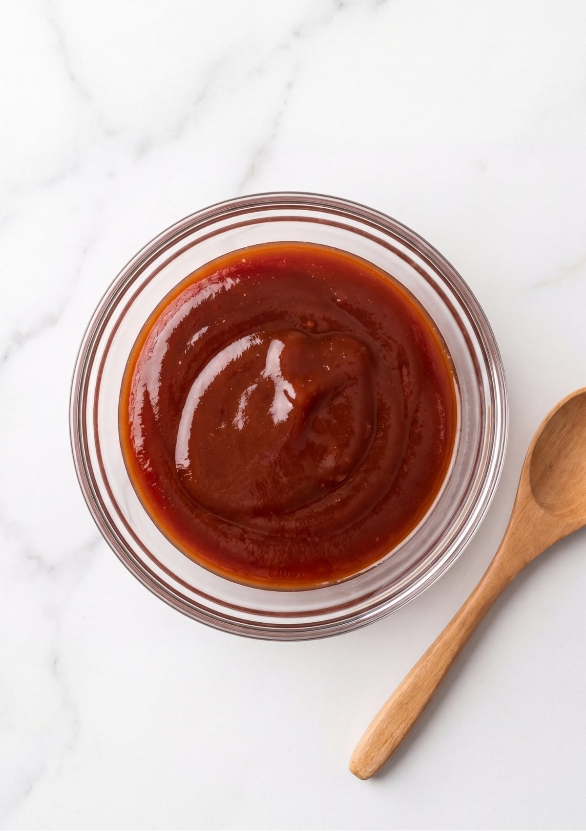 A smooth red meatloaf glaze in a small glass bowl next to a wooden spoon on a marble countertop