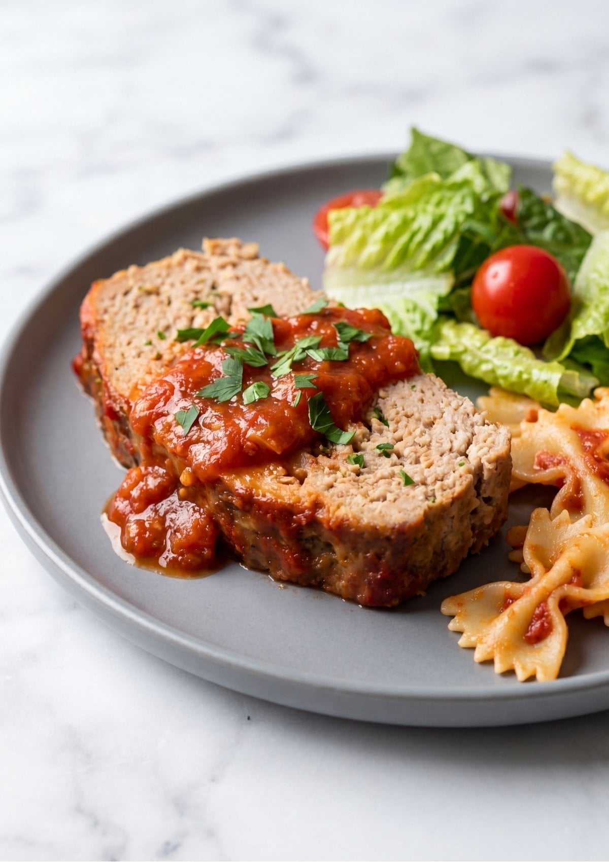 A gray plate featuring a slice of turkey meatloaf topped with bright red marinara sauce and chopped fresh parsley next to bow tie pasta and a green side salad with cherry tomatoes