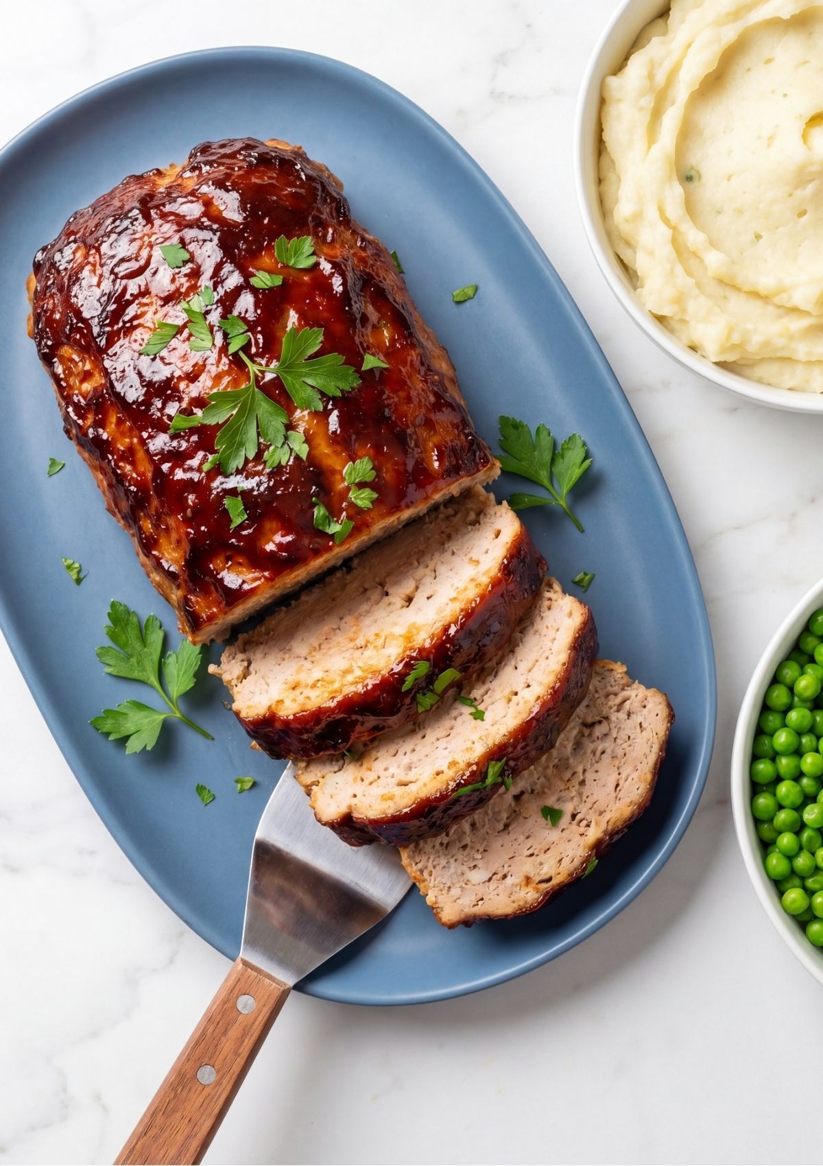 Slices of caramelized BBQ turkey meatloaf garnished with fresh parsley on a blue platter next to mashed potatoes.