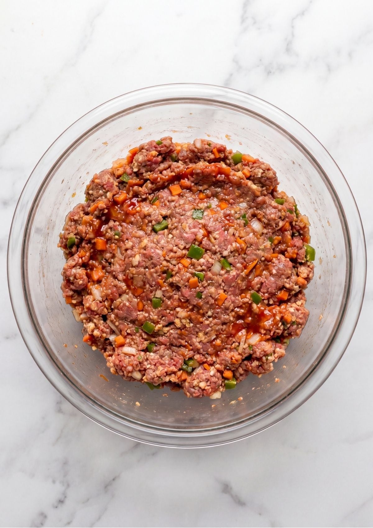 Ground meatloaf mixture combined with finely chopped sautéed vegetables and sauce inside a clear glass bowl.