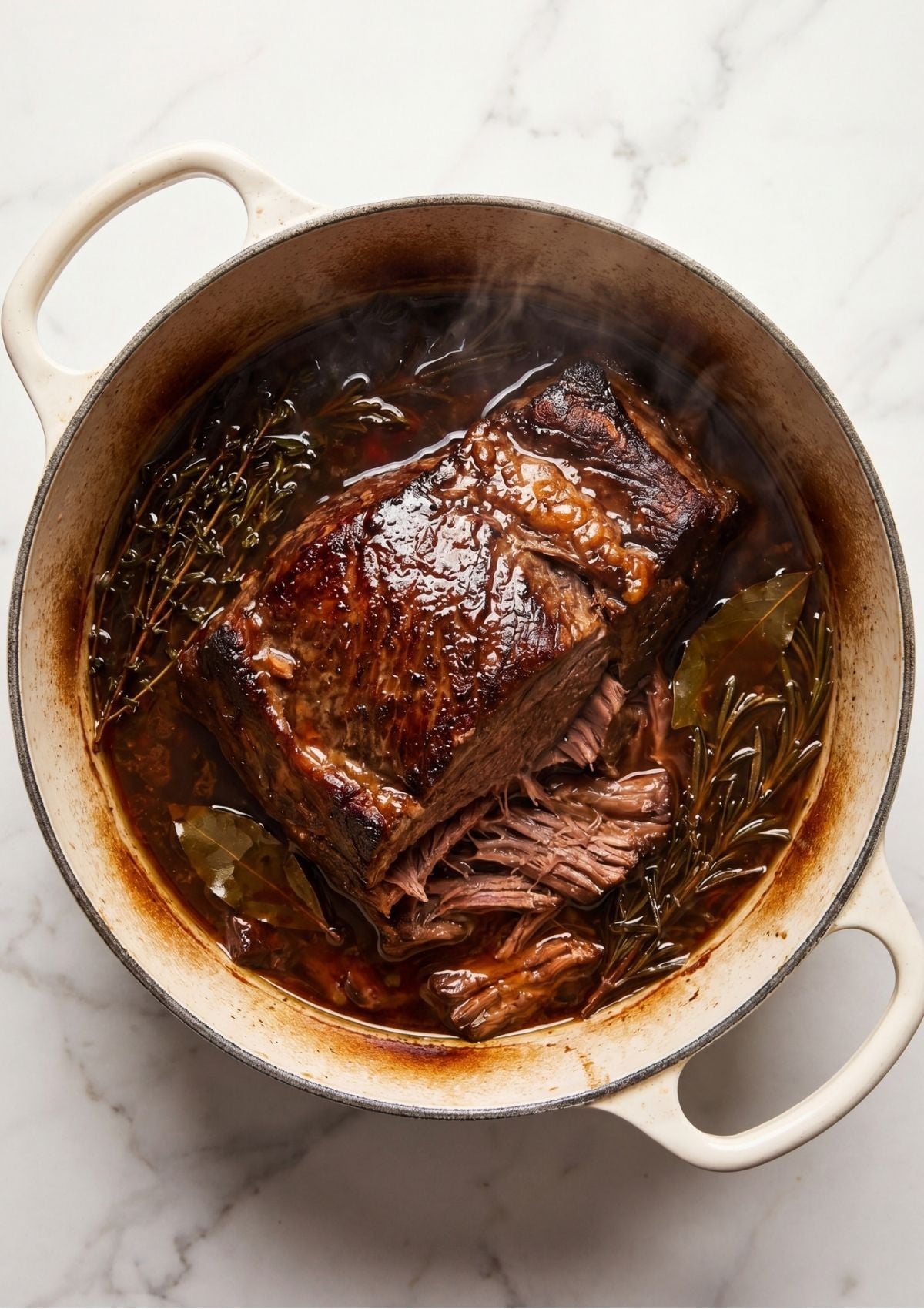 A fully cooked tender beef arm roast being pulled apart with a fork inside a Dutch oven with herbs and rich cooking juices.