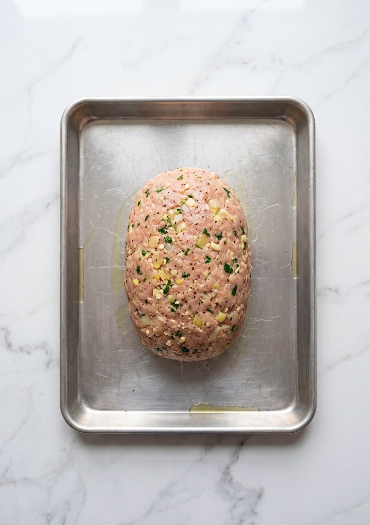 A raw ground turkey mixture formed into an oval loaf shape sitting on a silver rimmed baking sheet.