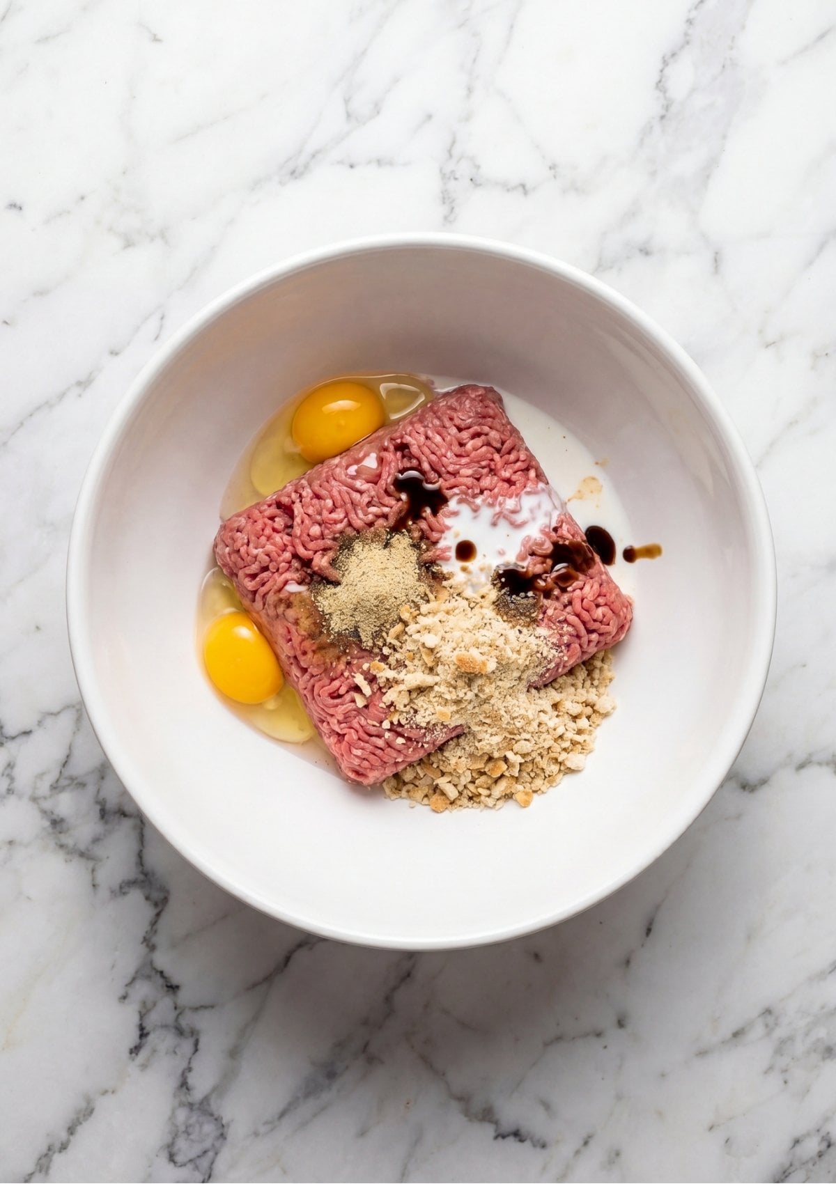 A white bowl containing raw ground beef, two cracked eggs, milk, and breadcrumbs ready to be mixed.