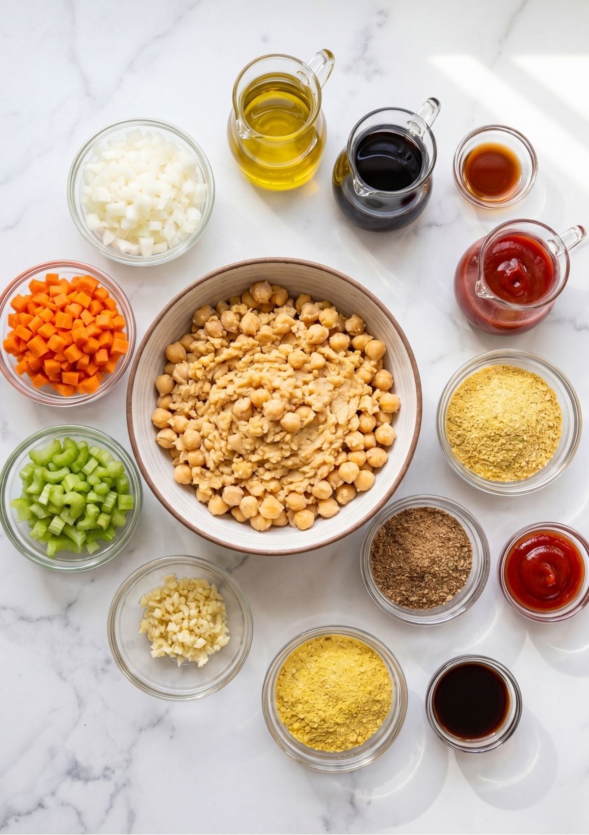 Flat lay of small glass bowls containing diced carrots, onion, celery, garlic, chickpeas, and spices for a vegan meatloaf recipe on a white marble background