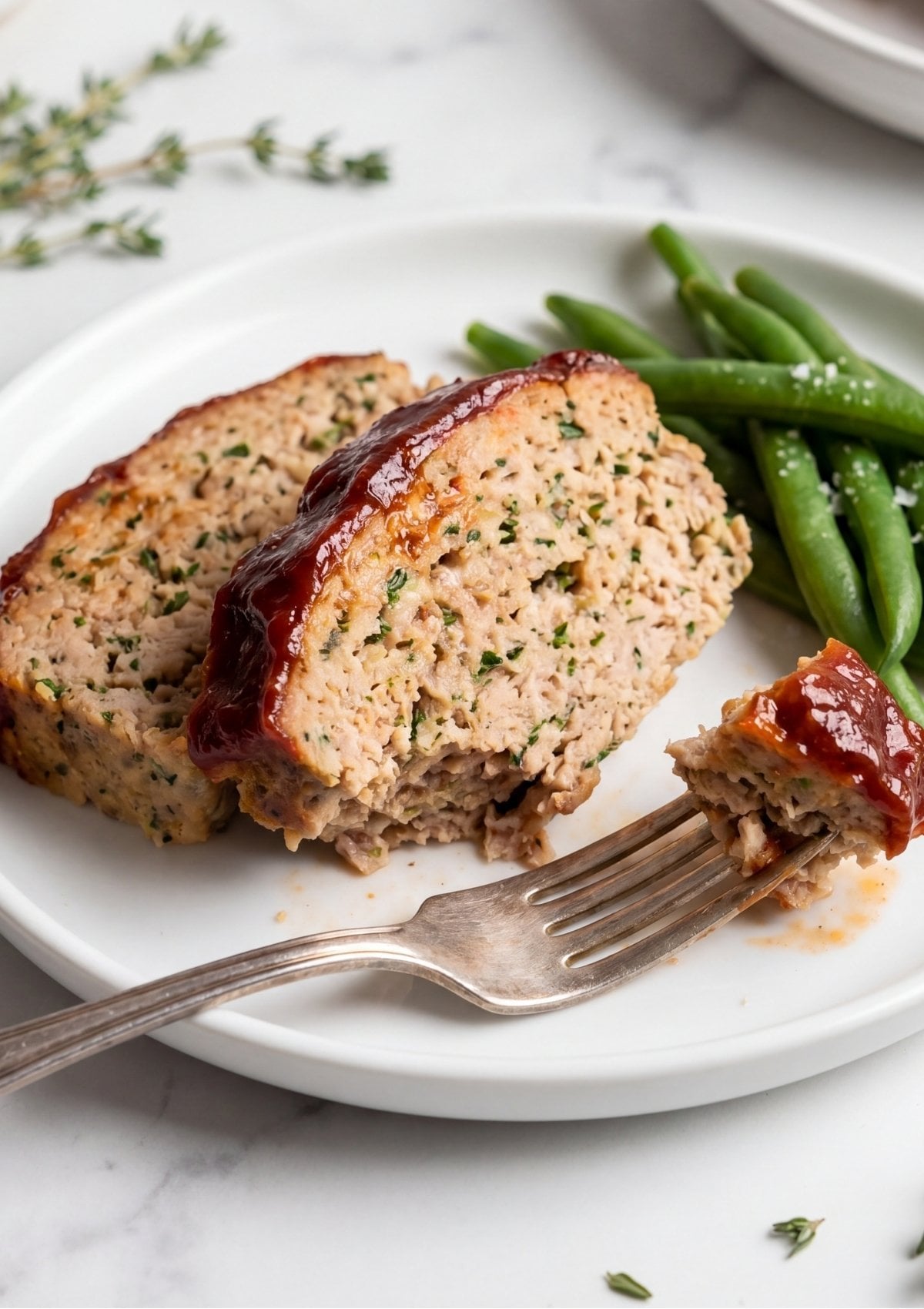 Two thick slices of glazed turkey meatloaf served on a white plate with a side of steamed green beans and a fork taking a bite.
