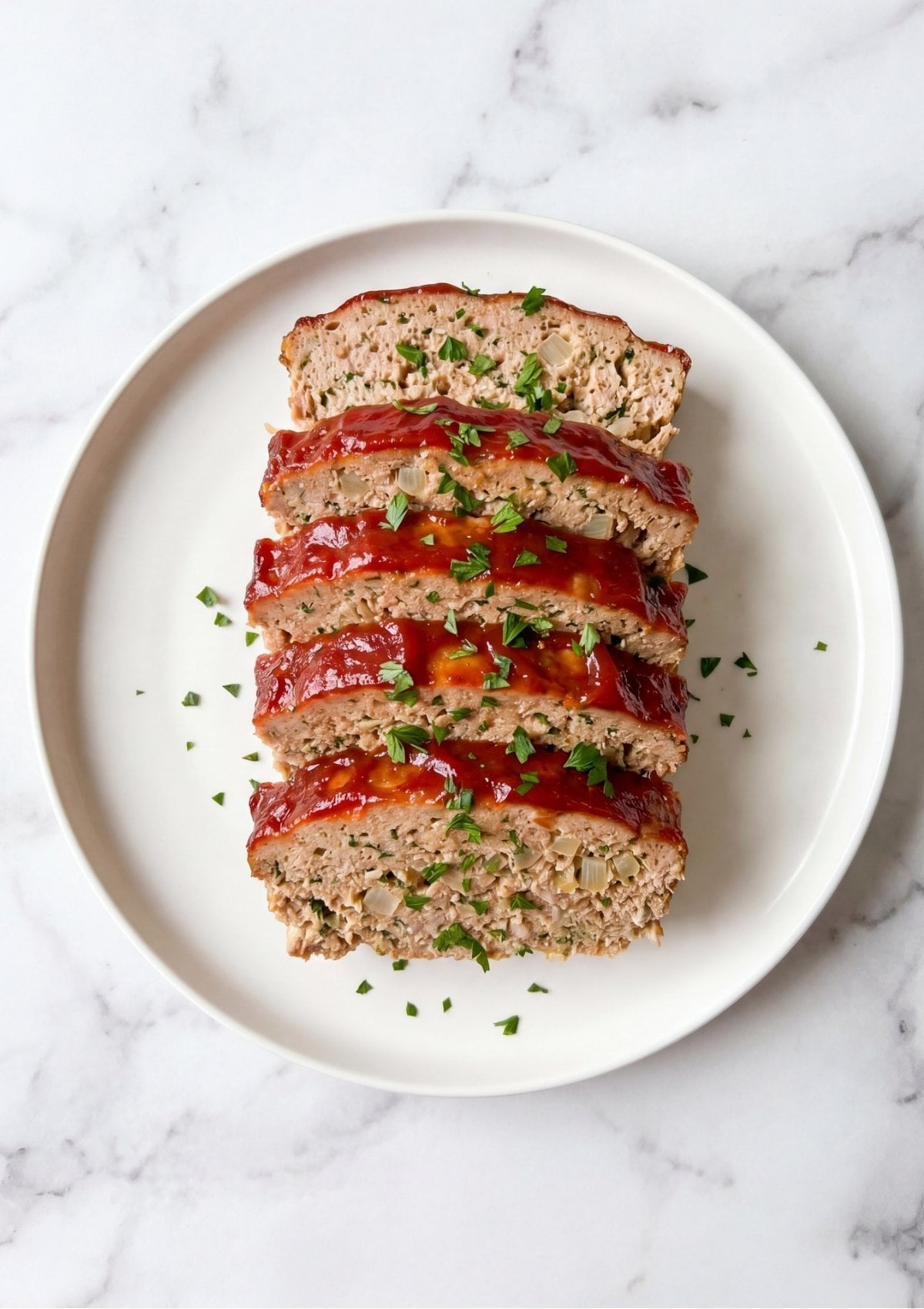 Overhead view of five thick slices of glazed turkey meatloaf arranged overlapping on a white ceramic serving plate, garnished with chopped green herbs
