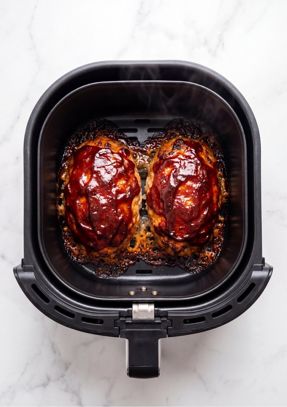 Overhead view of two fully cooked turkey meatloaves with a bubbly, caramelized ketchup glaze sitting inside an air fryer basket with steam rising