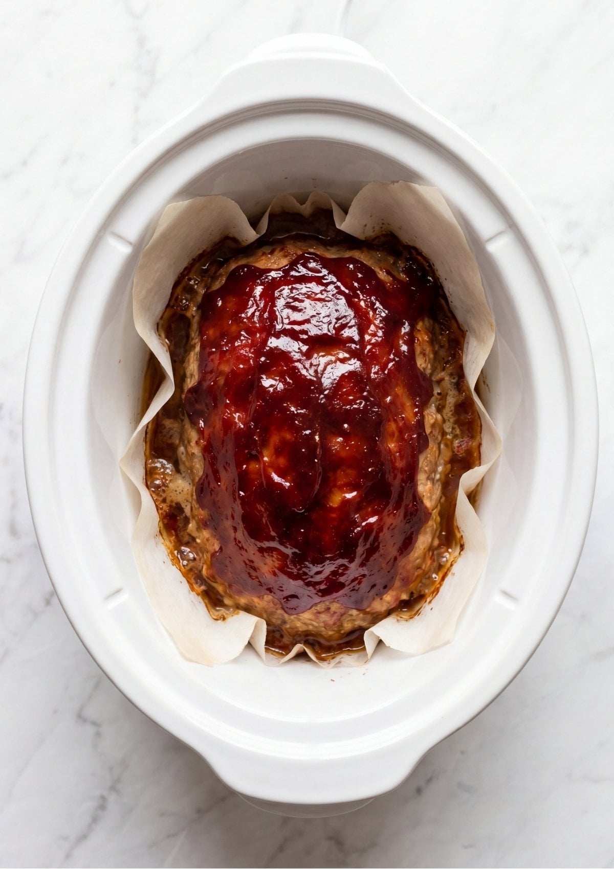 A whole turkey meatloaf cooking in a white crockpot lined with parchment paper topped with a generous layer of thick red ketchup glaze