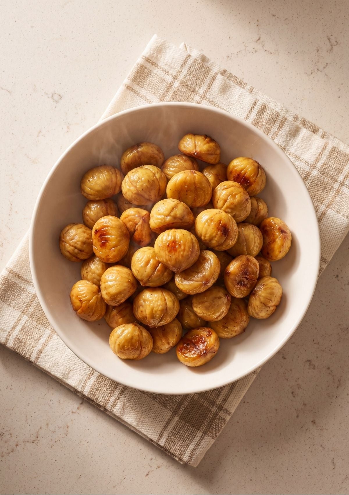 A white bowl filled to the brim with warm, golden-brown peeled chestnuts glistening with butter on a tan plaid napkin.