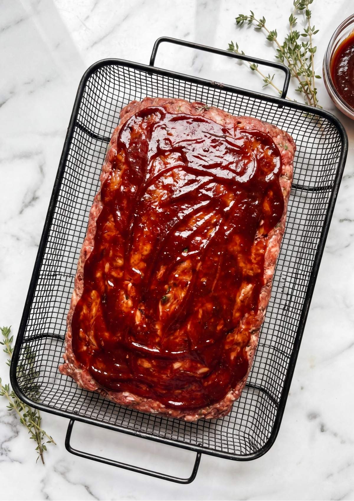 A raw meatloaf in a wire basket being brushed with a thick, dark red balsamic BBQ glaze, garnished with fresh thyme nearby.