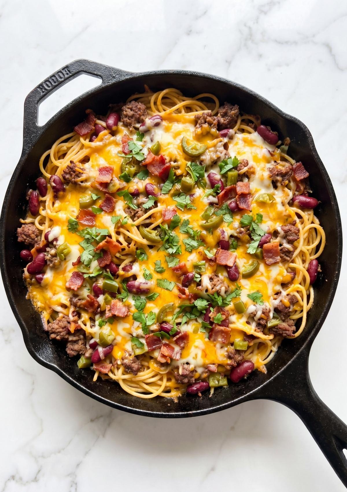 A completed Cowboy Spaghetti Casserole in a cast iron skillet topped with melted cheese, bacon, and cilantro.