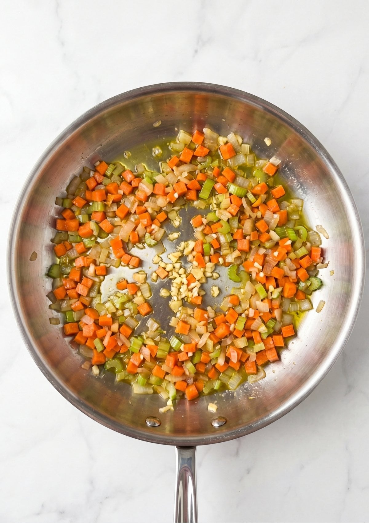 Finely diced carrots, celery, onion, and minced garlic sautéing in a silver skillet on a marble surface
