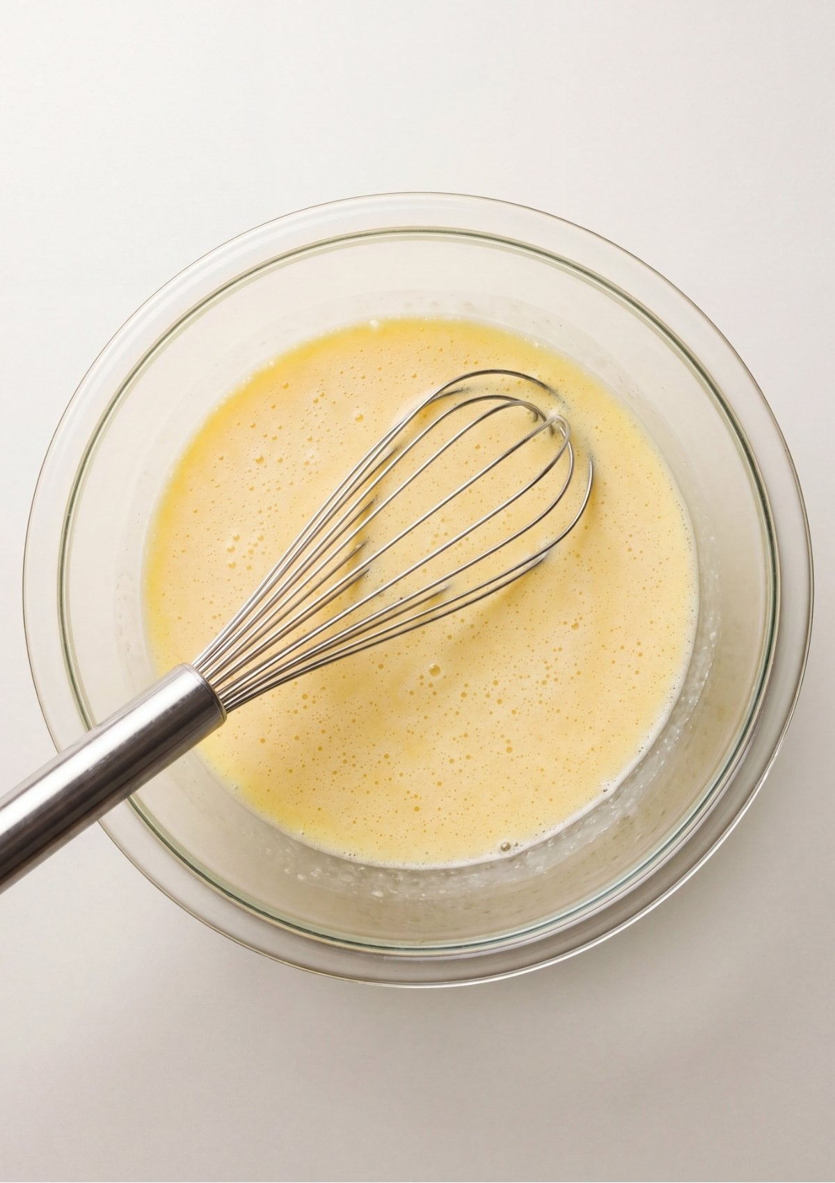 A metal whisk mixing egg and milk batter into a smooth and frothy mixture inside a clear glass bowl