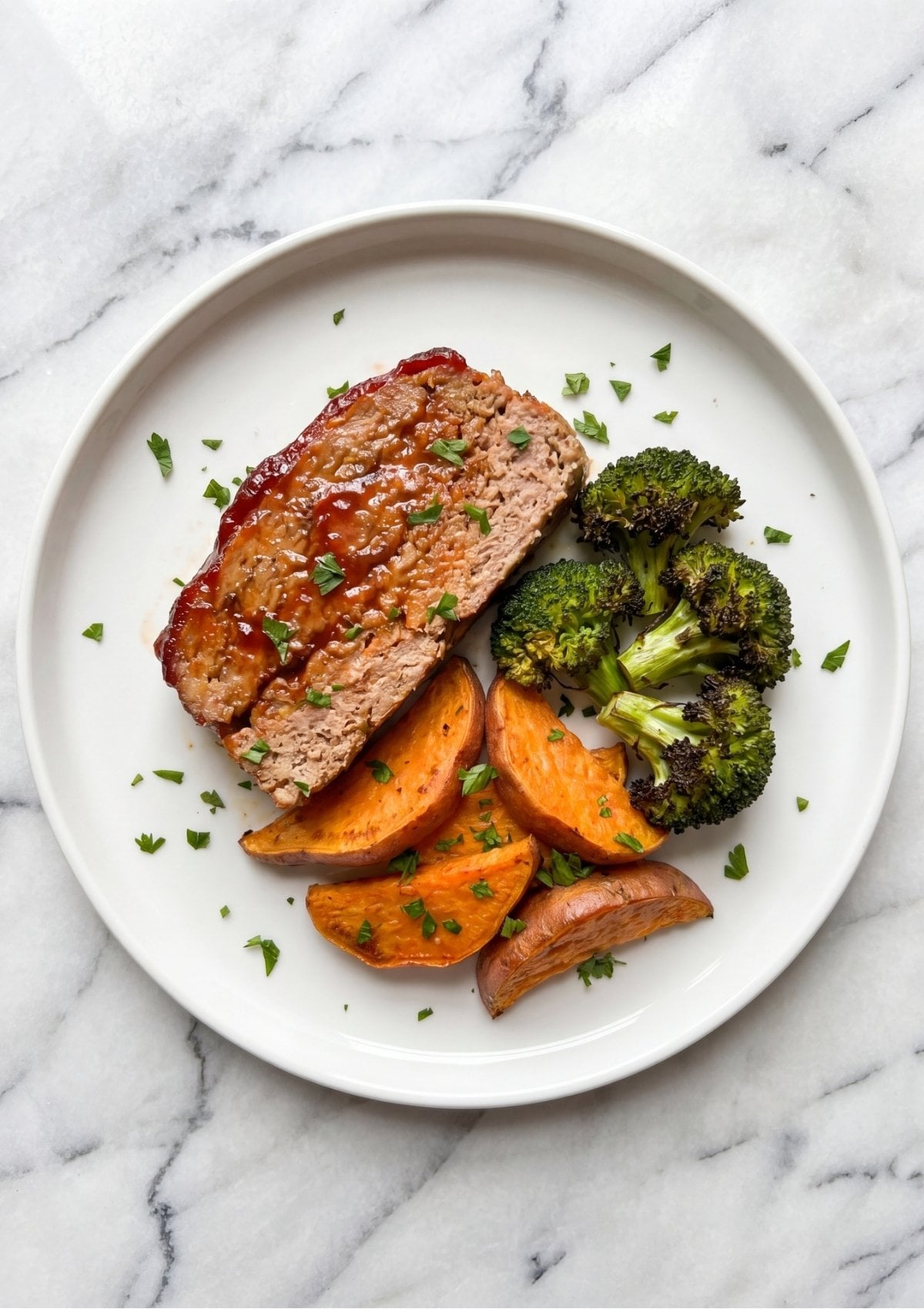 A single slice of glazed turkey meatloaf served on a round white plate alongside roasted broccoli florets and sweet potato wedges, sprinkled with fresh chopped parsley