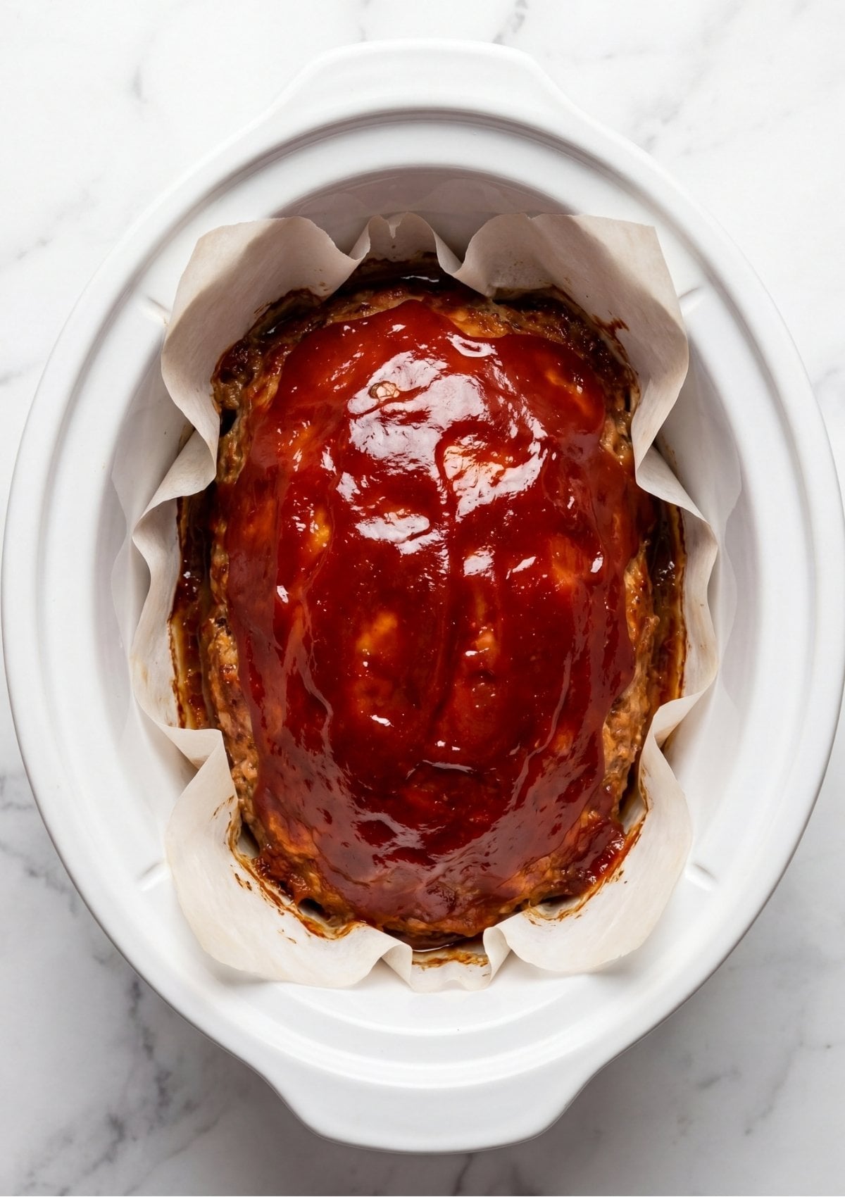 Top view of a turkey meatloaf covered in a smooth layer of red glaze inside a white crockpot lined with parchment paper