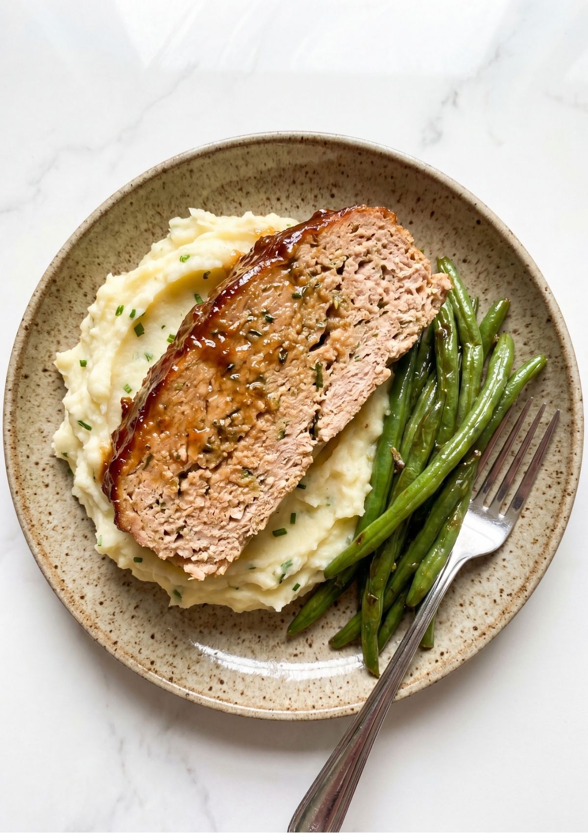 A thick slice of glazed turkey meatloaf served over a bed of creamy mashed potatoes with a side of green beans on a ceramic plate.