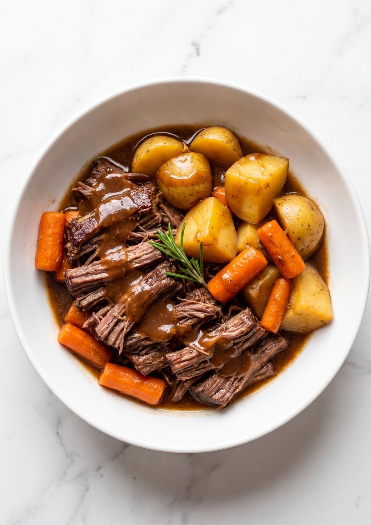 Sliced tender arm roast beef covered in savory brown gravy served with cooked carrots and potato chunks in a white bowl garnished with a sprig of rosemary.