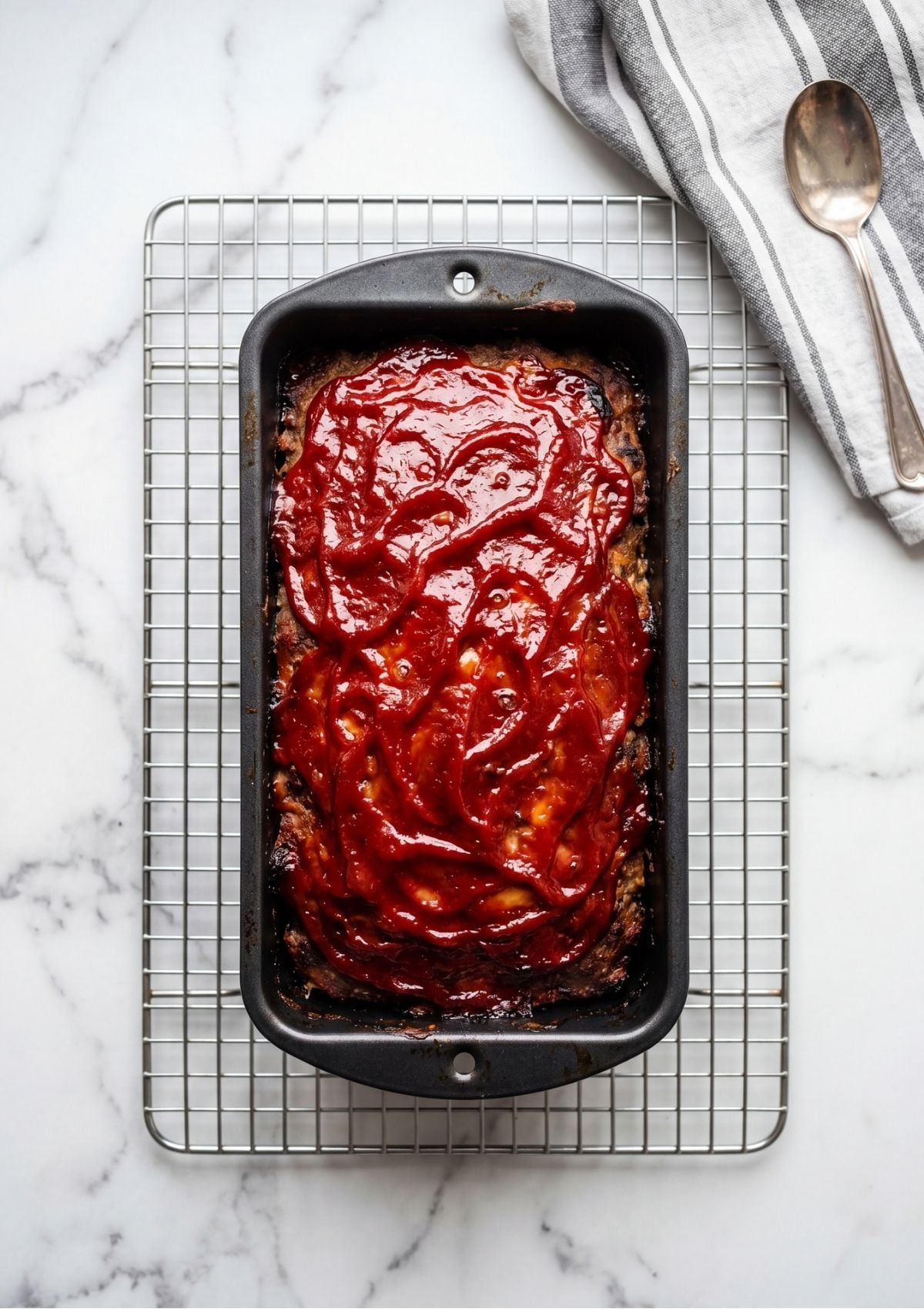 A glazed meatloaf resting in a pan on a wire cooling rack next to a striped kitchen towel.