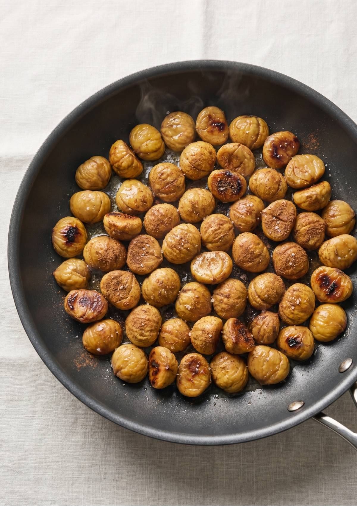 Steaming golden chestnuts lightly browned in a large black frying pan, sprinkled with ground cinnamon and coarse sea salt.