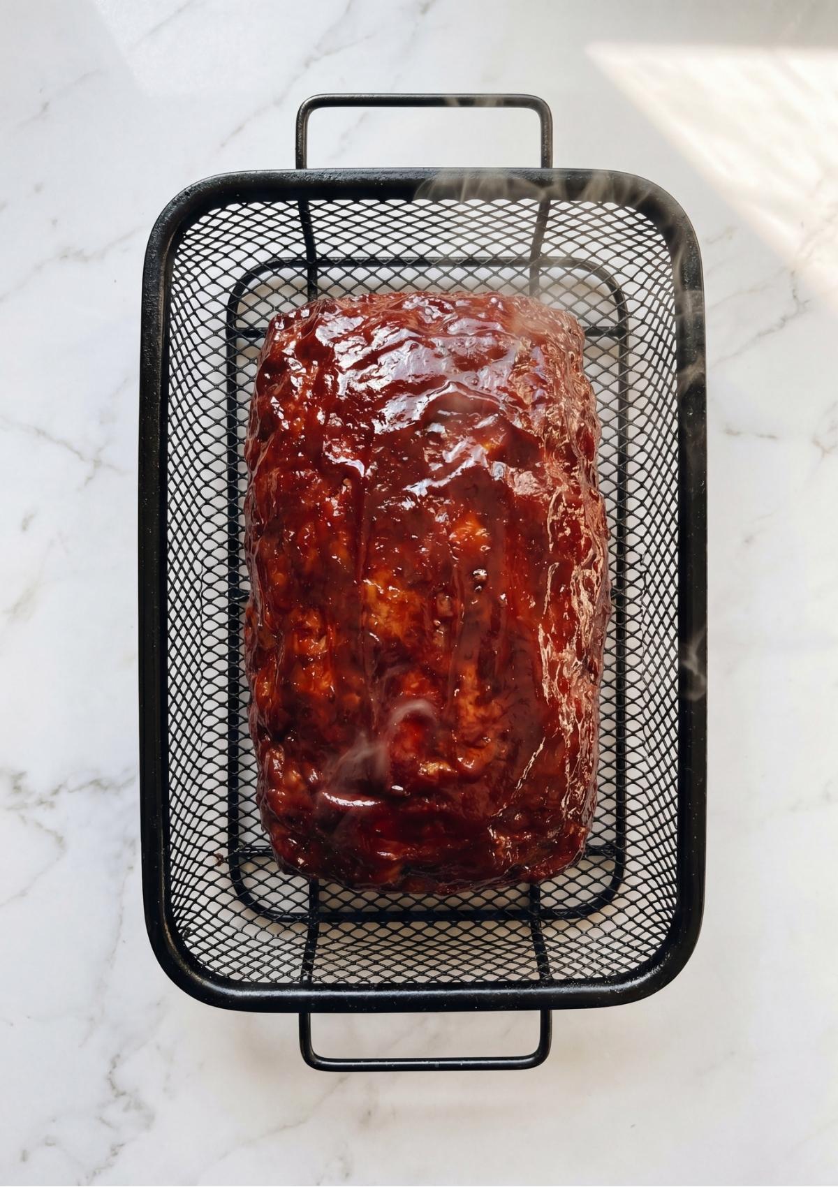 A fully cooked, steaming smoked meatloaf with a dark, caramelized glaze sitting in a black wire basket on a white marble countertop.