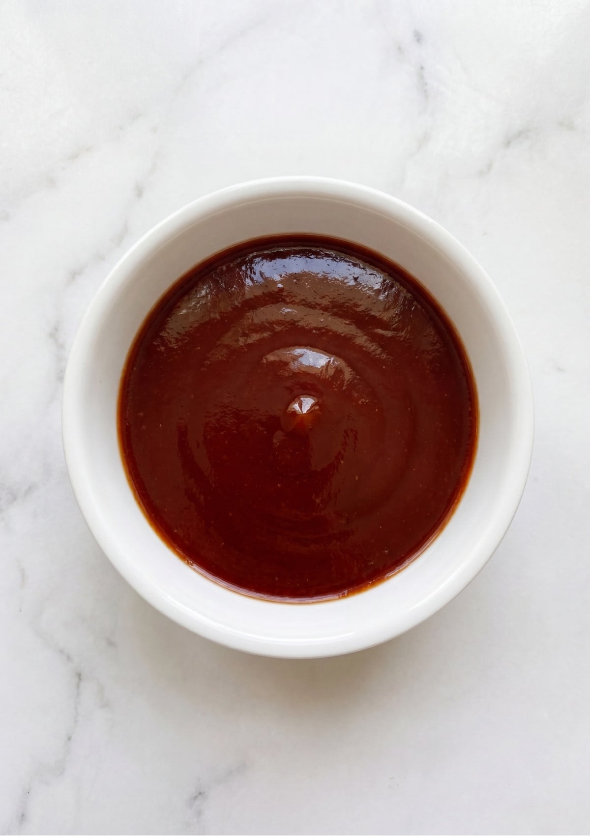 A white bowl filled with smooth, glossy reddish-brown BBQ glaze for topping a savory meatloaf.