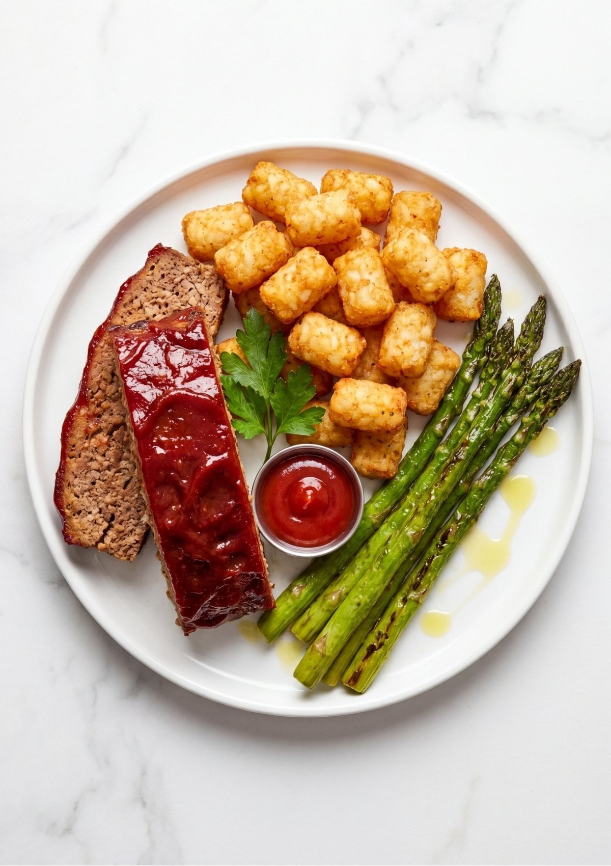 A white plate featuring two slices of glazed meatloaf, a pile of golden tater tots, and several green asparagus spears.