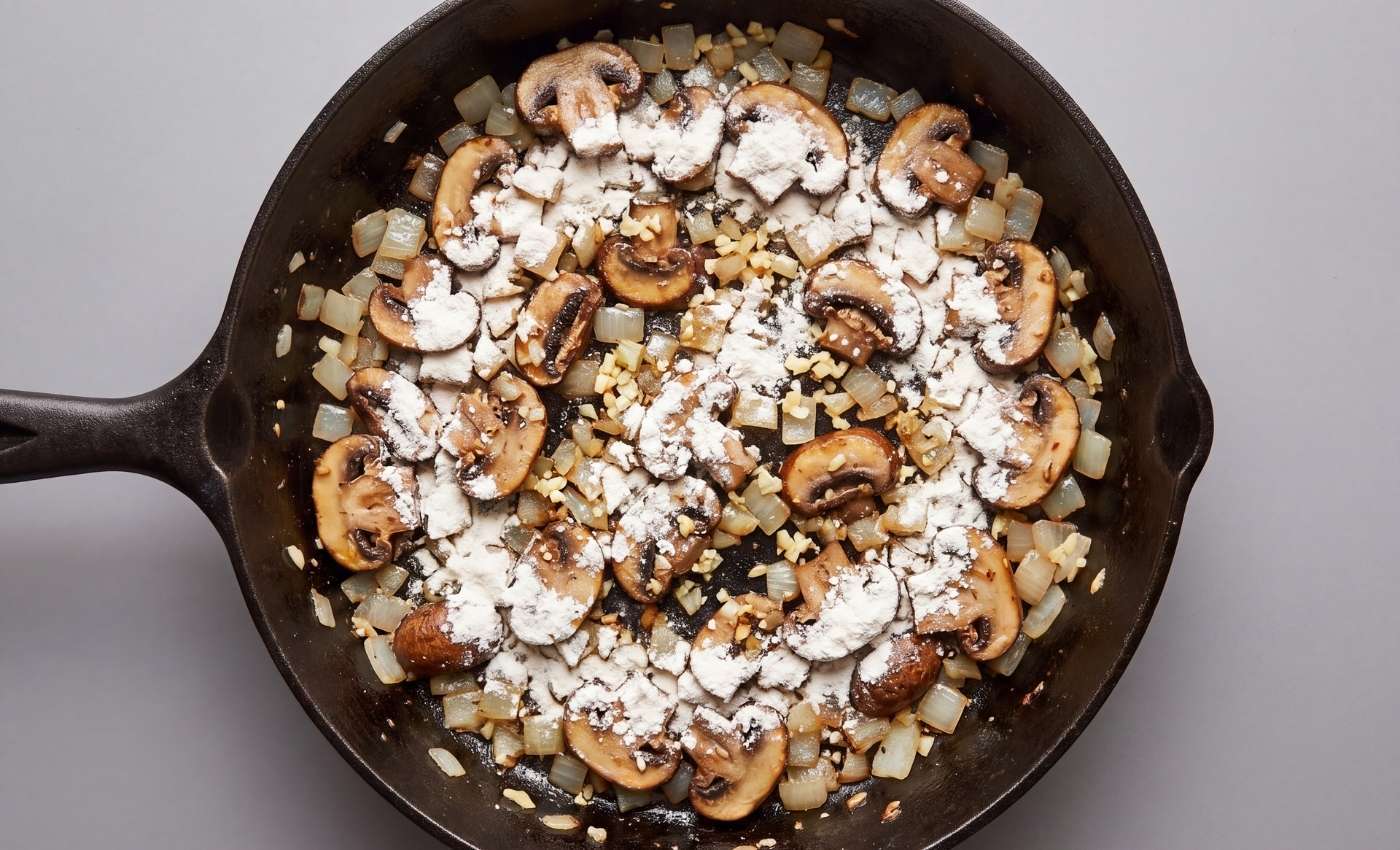 Sliced cremini mushrooms and onions cooking in a cast-iron skillet dusted with flour for beef stroganoff sauce