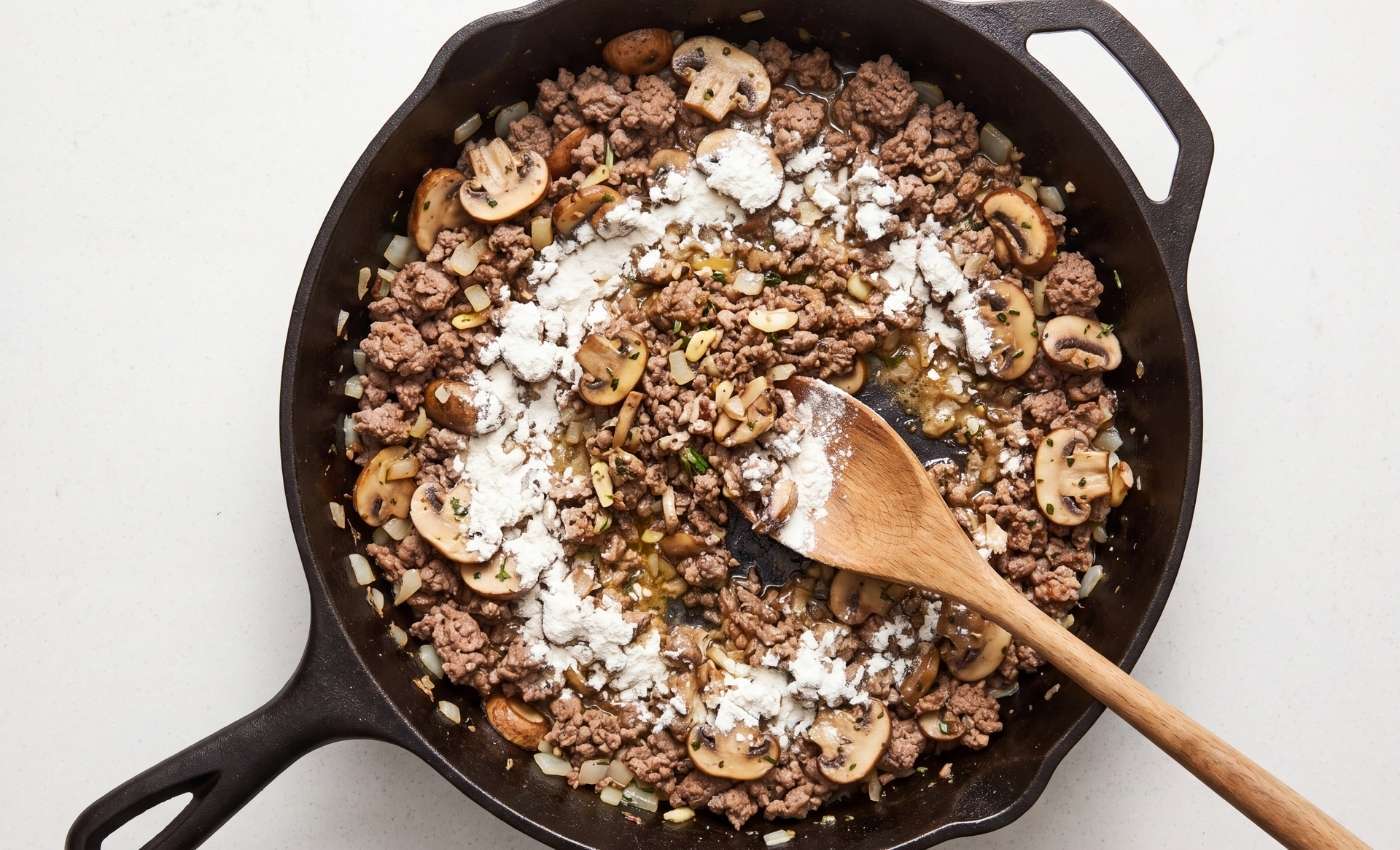 All-purpose flour sprinkled over the ground beef, mushrooms, onions, and garlic mixture in a cast-iron skillet