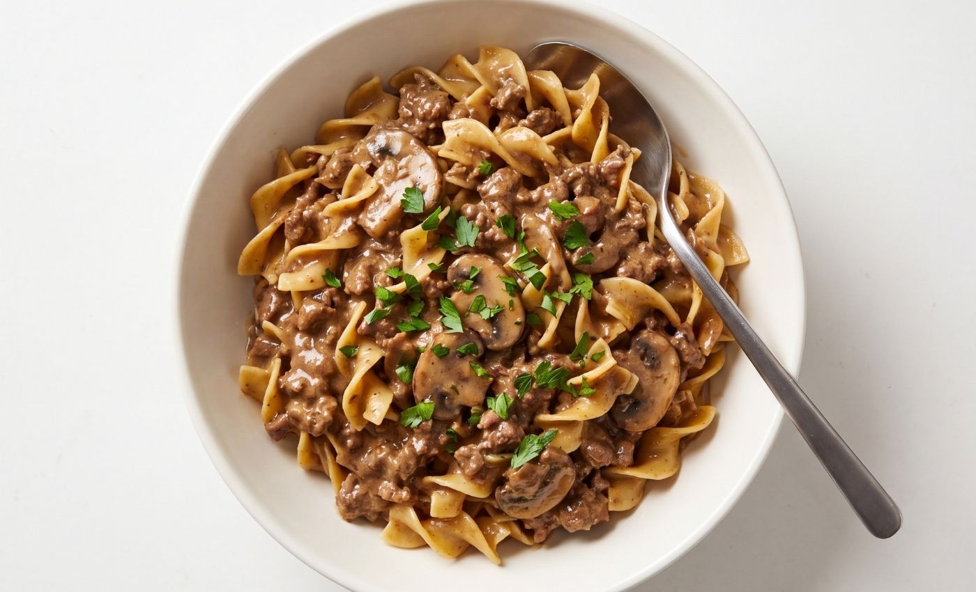A white bowl filled with ground beef stroganoff and creamy mushroom sauce over wide egg noodles with a spoon