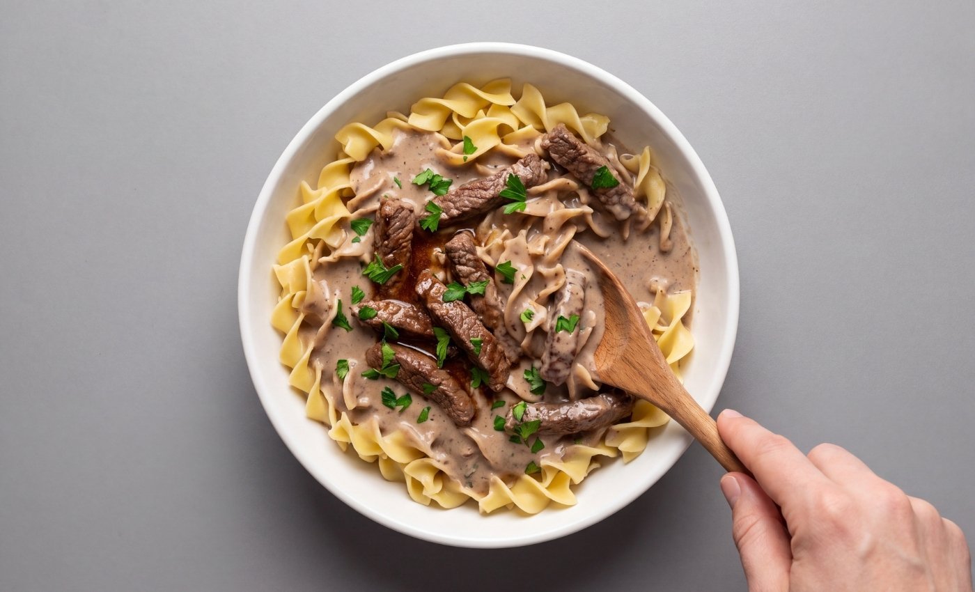 A serving of homemade beef stroganoff in a white bowl being stirred with a wooden spoon over egg noodles"